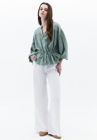 Camicia verde menta con bottoni e vita peplo, maniche oversize; indossata con pantaloni bianchi a gamba larga e scarpe in pelle intrecciata color nocciola. Tessuto texturizzato.