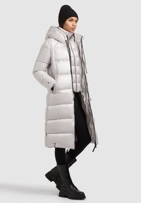 Hosszú ezüst színű steppelt pufi kabát kapucnival, fekete húzózsinórokkal és testhezálló belső réteggel. Fekete nadrággal és csizmával viselve.