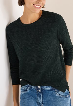 Dunkelgrüner, strukturierter Pullover mit langen Ärmeln und rundem Ausschnitt, kombiniert mit hellblauen Jeans aus Denim mit Knopfdetails.