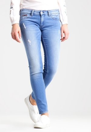 Jeans skinny de mezclilla azul claro con detalles desgastados, cinco bolsillos y cierre de botón, combinados con zapatos slip-on blancos.
