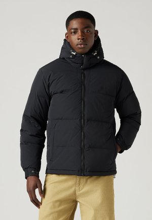 Must black puffer jakk, millel on kõrge kaelus, tõmbluku sulgemine ja reguleeritav kapuuts. Omab kahte rinna-taskut ja elastseid mansette. Sile tekstuur.