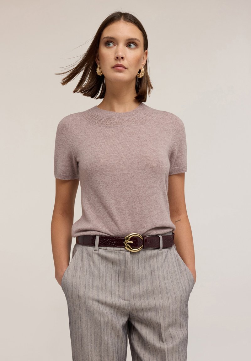 Svetle růžový pletený svetr s krátkým rukávem a kulatým výstřihem, doplněný šedými pruhovanými kalhotami a vínovým páskem se zlatou kulatou sponou.