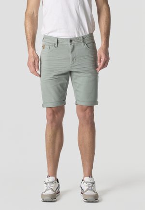 Hombre con pantalones cortos de dobladillo enrollado de color gris claro, camiseta blanca y zapatillas blancas, de pie frente a un fondo liso.