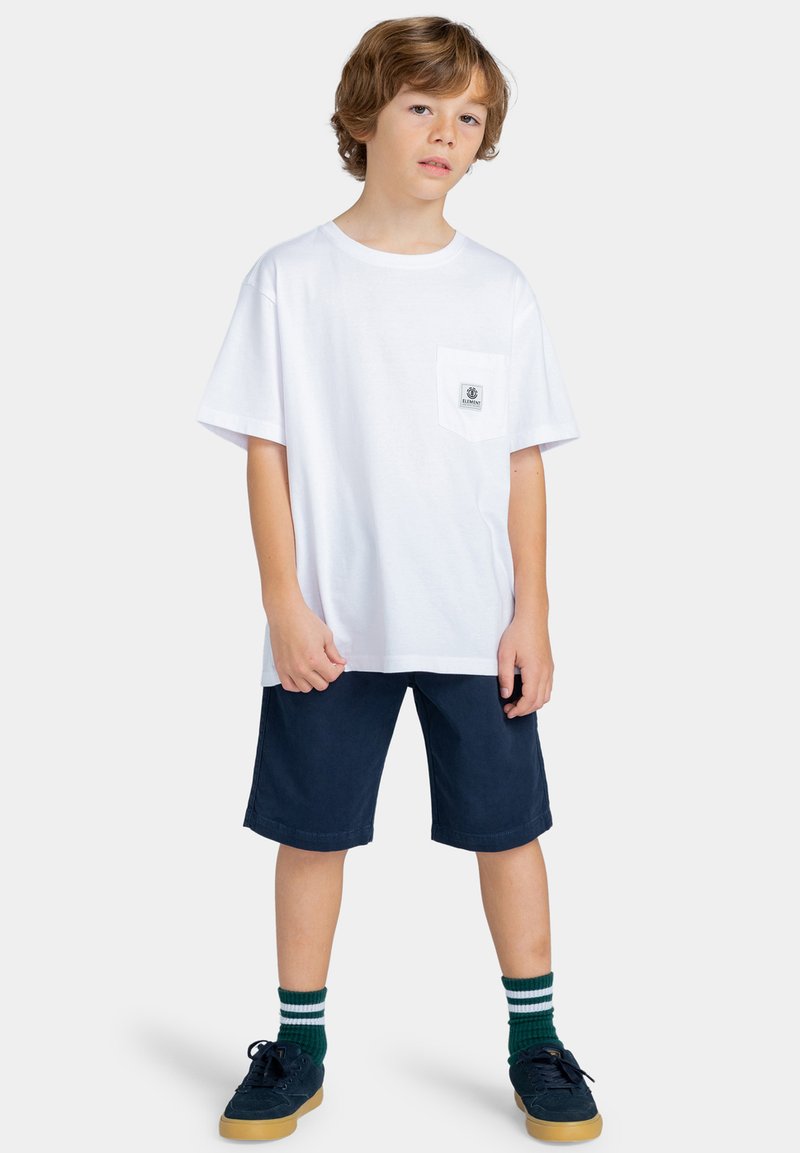 Witte katoenen T-shirt met een borstzak, gecombineerd met donkerblauwe shorts. Kind draagt groen-witte gestreepte sokken en donkere sneakers.
