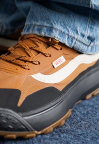 Vans sportbačiai, pagaminti iš rudos ir juodos medžiagos, turi baltą bangos raštą, tekstūruotą paviršių ir iškilų juodą pado dizainą su giliu padu.