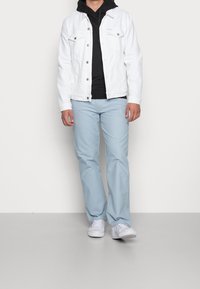 Persona vestida con una chaqueta de mezclilla blanca sobre una sudadera negra, pantalones azul claro y zapatillas blancas, de pie sobre un suelo gris con un fondo blanco.