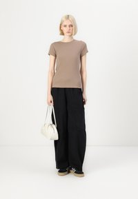 Hnědý žebrovaný top s krátkým rukávem v kombinaci s oversized černými kalhotami. Modelka drží bílou kabelku a má na sobě černé tenisky se světlou podrážkou.