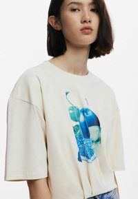 T-shirt en coton couleur crème avec des manches courtes et un grand imprimé graphique bleu et vert représentant des fruits et des feuilles stylisés sur le devant.