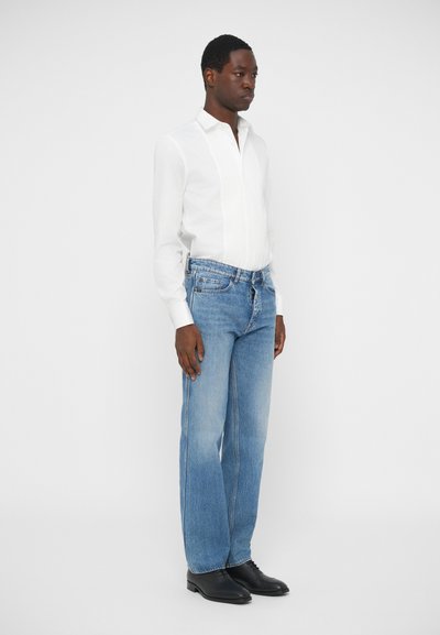 Bela srajca z dolgimi rokavi in vertikalnimi črtami, v kombinaciji s svetlo modrimi ravnimi kavbojkami in črnimi čipkastimi čevlji. Enobarvna ozadje.