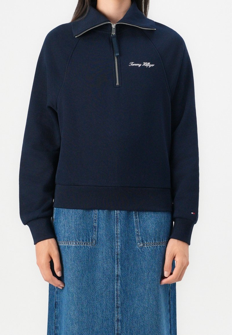 Femme portant un sweat-shirt zippé bleu marine avec le logo "Tommy Hilfiger" et une jupe en denim bleu.