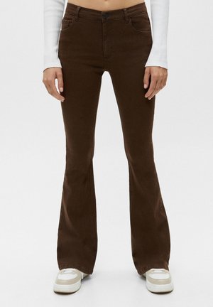 Braune hoch-taillierte, ausgestellte Hose getragen mit weißen Sneakers und einem weißen langärmeligen Oberteil, von der Taille bis zu den Füßen vor einfachem Hintergrund gezeigt.