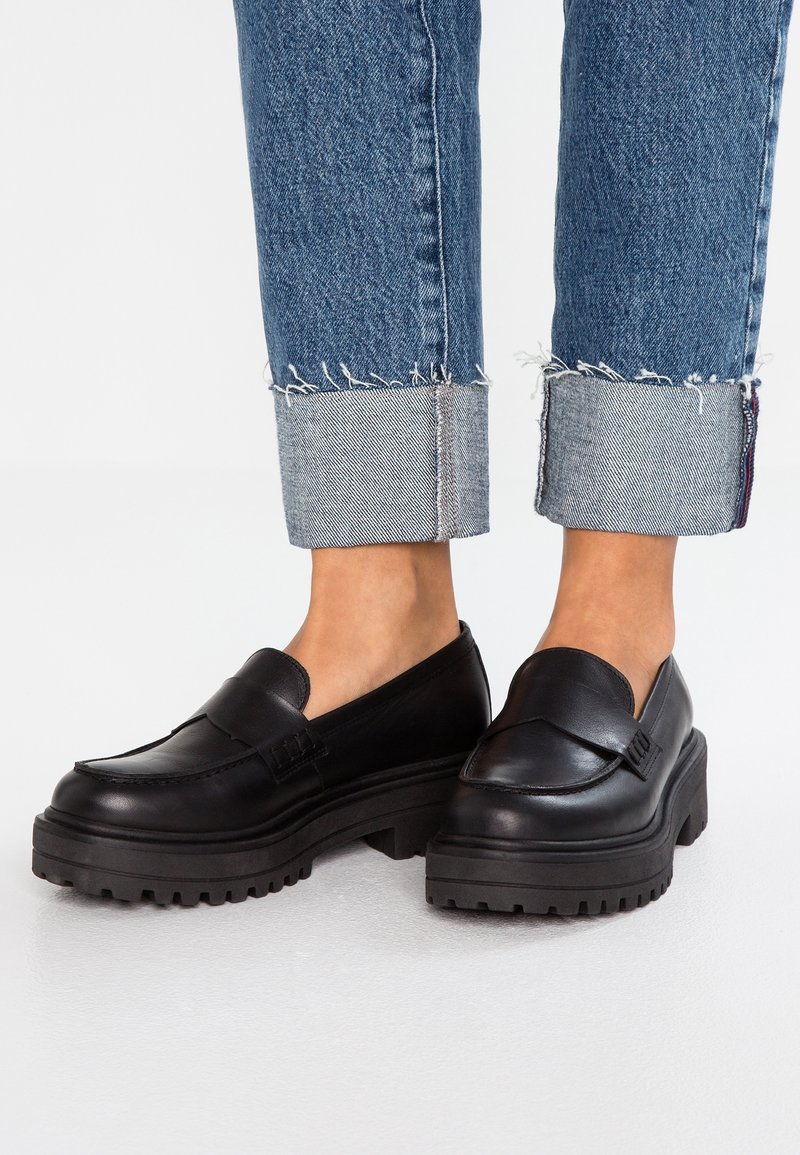 Mocassins à plateforme épaisse noirs portés avec un jean en denim bleu retroussé sur fond blanc.