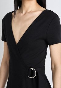 Czarna krótka sukienka kopertowa z głębokim dekoltem w serek i srebrnym zapięciem w talii, noszona przez osobę z prostymi, ciemnymi włosami.