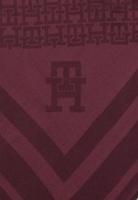 Burgunderrød stof med et zigzagmønster, der har mørke nuancer og et gentaget logo-design i midten. Blødt, tekstureret materiale.