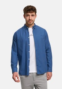 Nuori mies, jolla on kiharat hiukset, sininen nappipaita valkoisen painokuvioidun t-paidan päällä ja vaaleanharmaat housut, seisoo yksinkertaisen taustan edessä.