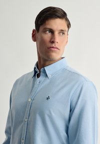 Camisa de botonadura clásica de algodón en azul claro con mangas largas, que presenta un logotipo bordado en negro en el pecho y un diseño de cuello estándar.