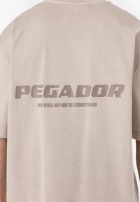 Bežna bombažna majica z velikim, poudarjenim napisom "PEGADOR" na hrbtni strani, pod katerim se nahaja stavek "CERTIFIED AUTHENTIC STREETWEAR".