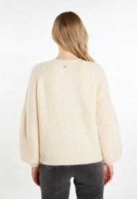 Donna con lunghi capelli biondi che indossa un maglione a coste color crema e jeans grigio scuro, vista di spalle su uno sfondo bianco.