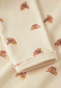 Tessuto di cotone con una texture a coste, con sfondo beige e piccoli motivi a croissant in tonalità marrone e oro, con bordo ondulato.