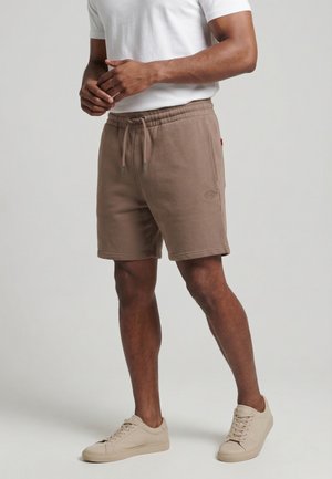 Homme portant un short beige à cordon, un t-shirt blanc et des baskets beiges, debout devant un fond clair uni.