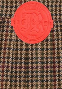 Bruin en beige houndstooth stof met een rode, gestructureerde, ronde patch met de tekst "Richmond" en 1984, voorzien van ingewikkelde stikdetails.