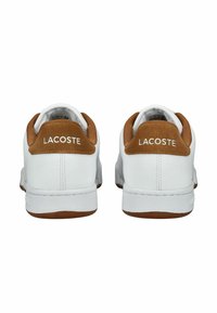 Bílé kožené tenisky s hnědými semišovými akcenty na patě, s plochou gumovou podrážkou a logem "LACOSTE" na zadní straně.