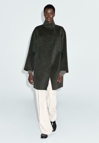 Model v temně šedém oversized vlněném kabátu s vysokým límcem, světlých béžových širokých kalhotách a černých botách, kráčející na jednolitém světlém pozadí.