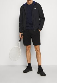 Black lightweight jacket with blue accents, navy t-shirt, black shorts, and black athletic shoes. Holding a white tennis racket.