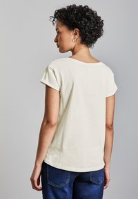 Krémové bavlněné tričko s kulatým výstřihem a krátkými ohrnutými rukávy, v kombinaci s tmavě modrými džíny. Hladká textura, ležérní střih.