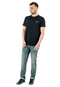 T-shirt di cotone nera con collo rotondo e maniche corte, caratterizzata da un piccolo emblema sul lato sinistro del petto; abbinata a jeans blu chiaro e sneaker nere.