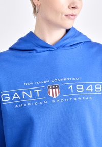 Hanorac albastru dintr-un material moale, având o grafică frontală cu "GANT 1949," un simbol și textul "AMERICAN SPORTSWEAR." Designul hanoracului include șnur.