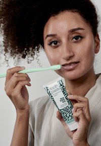 Tannrør med teal og svart botanisk design, merket "tea tree oil." Brukeren holder en mintgrønn tannbørste og påfører tannkrem.