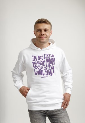 Mann trägt weißen Kapuzenpullover mit lila Text "I'm not like a regular mom, I'm a cool mom" aus Mean Girls, steht vor schlichtem Hintergrund.