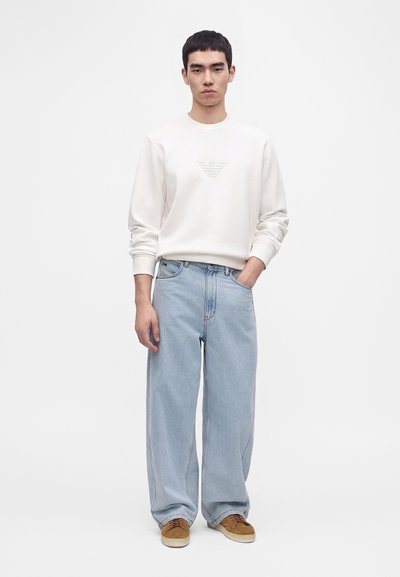 Jeune homme aux cheveux courts et foncés portant une chemise blanche à manches longues, un jean large bleu clair et des baskets marron, debout avec une main dans la poche.