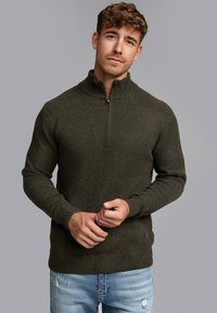 Sudadera de cremallera verde oscuro con cuello alto, puños y dobladillo acanalados, de tejido de punto con textura. Llevada con vaqueros claros desgastados.