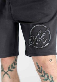 Pantaloni shorts neri in tessuto morbido presentano un logo ricamato in bianco ben visibile. La pelle con tatuaggi di foglie verdi è parzialmente visibile.