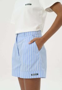 Jasnoniebieskie szorty w pasy z białymi pionowymi liniami, z przednimi kieszeniami i czarną naszywką z logo na dole. Wykonane z gładkiego materiału.