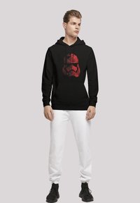 Juodas puloveris su gobtuvu, ant kurio priekio pavaizduota didelė raudona geometrinė stormtrooper dizaino iliustracija, derinama su baltais sportiniais kelniais ir juodais sportbačiais.