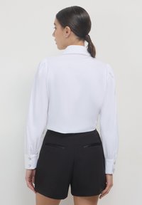 Weißes Langarmbluse mit strukturiertem Kragen und Knopfakzenten, kombiniert mit schwarzen Shorts mit Gesäßtaschen und glatter Oberfläche.