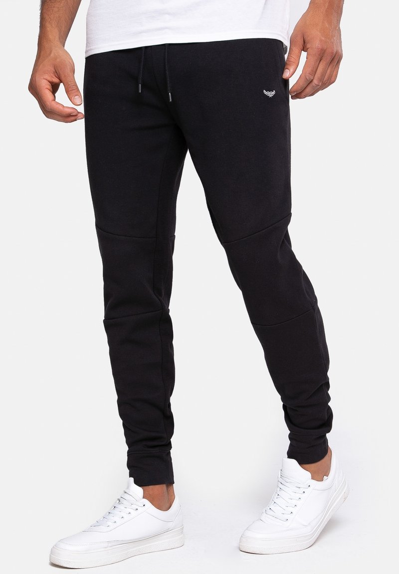 Un homme porte un pantalon de jogging noir fuselé avec une taille à cordon et des baskets blanches, debout devant un fond blanc uni.
