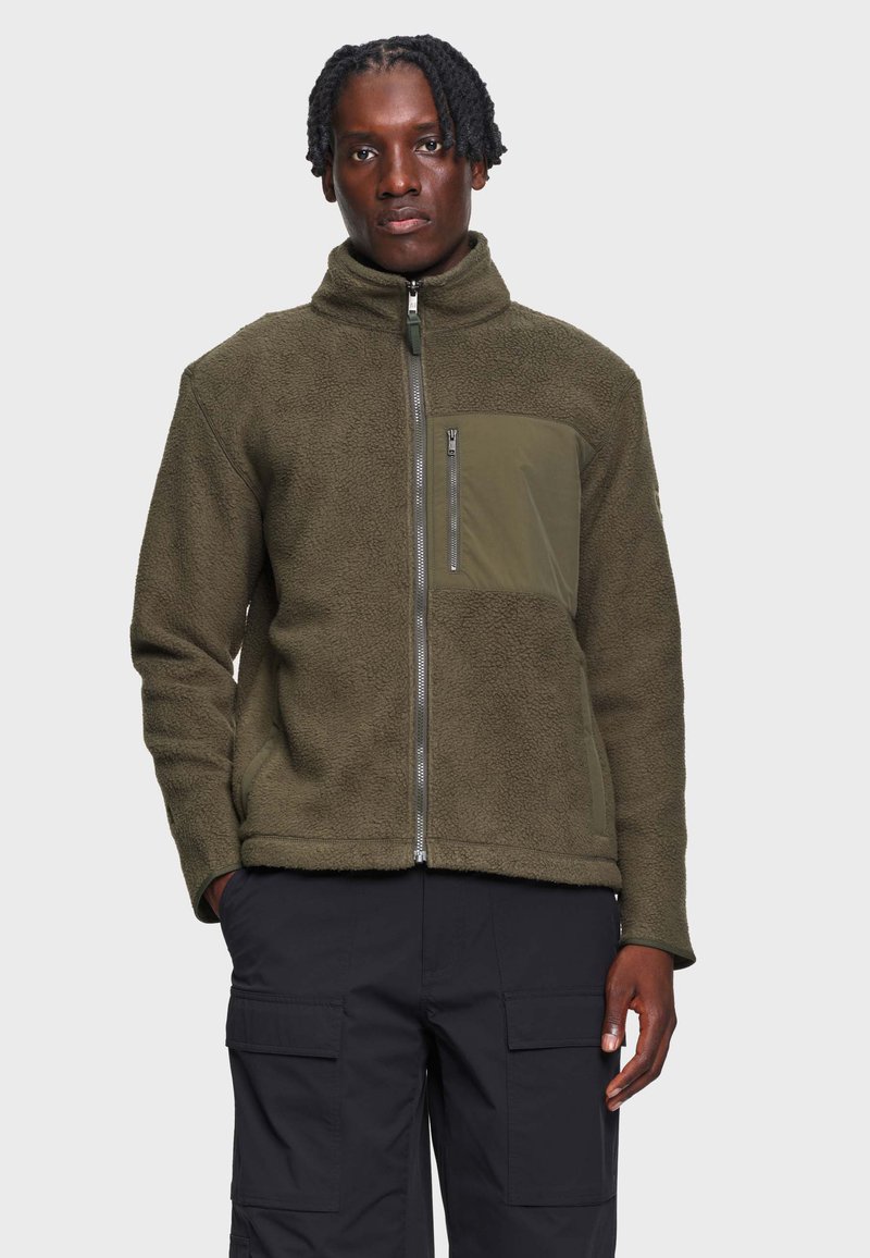 Homme aux cheveux tressés portant une veste polaire vert olive avec une poche poitrine zippée et un pantalon cargo noir, debout devant un fond uni.