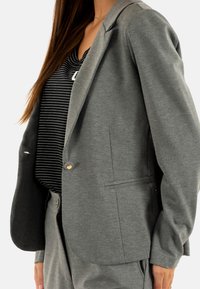 Blazer gris hecho de tela suave con solapas de muesca, dos bolsillos frontales y un cierre de un solo botón dorado. Llevado sobre una camiseta de rayas.