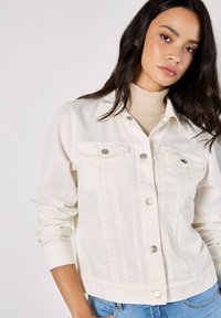 Chaqueta de mezclilla beige claro con botones a presión plateados, dos bolsillos en el pecho, puños acanalados y detalles de costura vertical. Material de algodón suave.