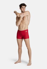 Rode zwembroek van stretchstof, met een logo op de taille. Het model neemt een atletische houding aan, waardoor de pasvorm en het ontwerp zichtbaar zijn.