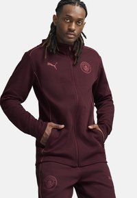 Giacca zip-up borgogna con accenti rosa, tasche laterali e logo del Manchester City. Indossata con pantaloni borgogna abbinati, in tessuto misto cotone.