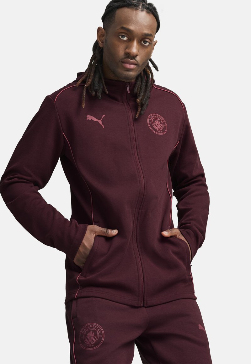 Bordo zip-up jaciņš ar rozā akcentiem, sānu kabatām un Manchester City logo. Valkāts kopā ar atbilstošām bordo biksēm, kokvilnas maisījuma audums.