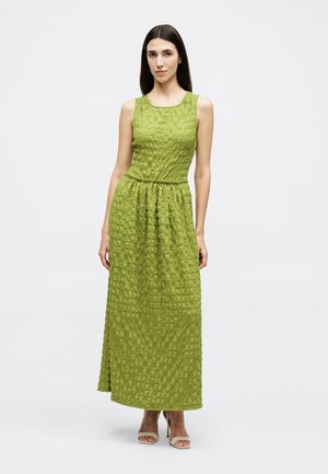 Γυναίκα με ίσια σκούρα μαλλιά που φοράει αμάνικο μάξι φόρεμα σε υφή λάιμ πράσινου χρώματος και μπεζ παπούτσια ανοιχτά στα δάχτυλα, στέκεται μπροστά σε λευκό φόντο.