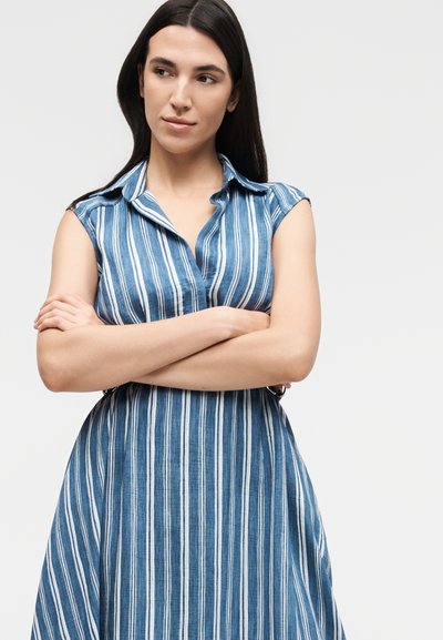 Γυναίκα με μακριά σκουρόχρωμα μαλλιά που φοράει μπλε και λευκό κατακόρυφο ριγέ αμάνικο φόρεμα, με σταυρωμένα τα χέρια, κοιτάζοντας προς τα πλάγια.