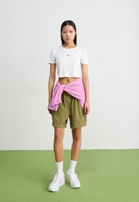 T-shirt bianca a maniche corte, pantaloni corti in velluto a coste verde, camicia a quadri rosa legata in vita, sneakers bianche con dettagli rosa, posate su un pavimento verde.