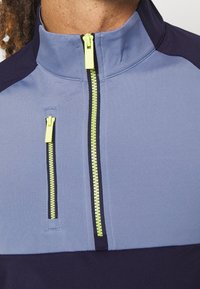 Gros plan d'une veste bleu et marine avec deux fermetures éclair verticales jaune vif, une au centre et une sur la poche poitrine gauche, portée par une personne.